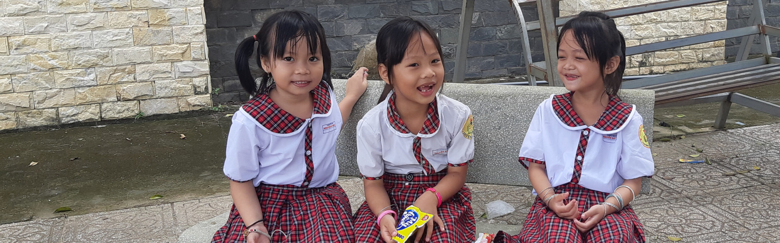 Three young school girls sitting outside their classroom smiling and laughing together in their school uniforms